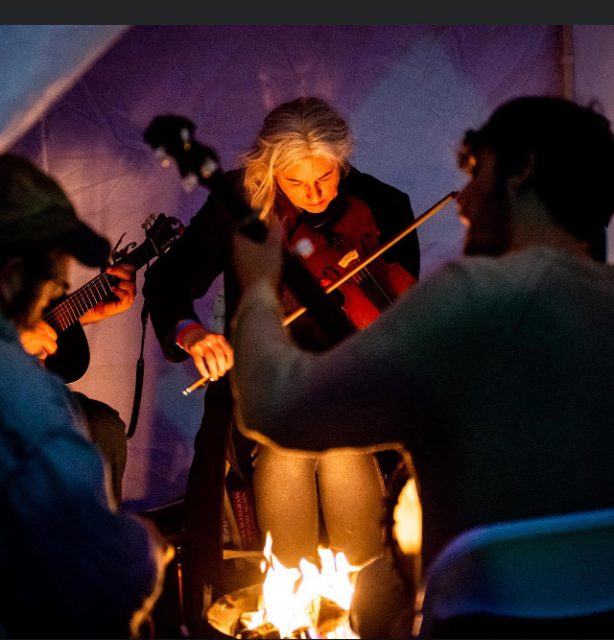 Woman playing fiddle in old-time music group at night around campfire